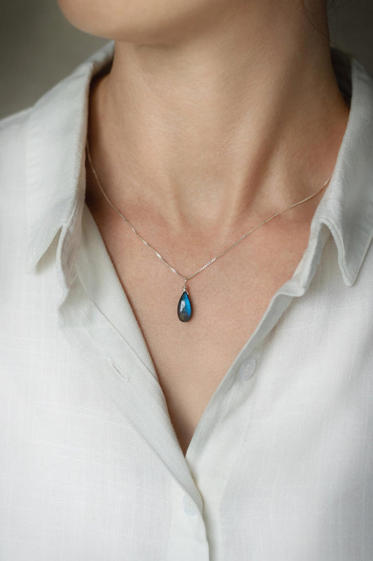 A handcrafted Labradorite teardrop necklace pendant showing a stunning blue and green iridescent flash, on a sterling silver chain, worn by a person in a white shirt.