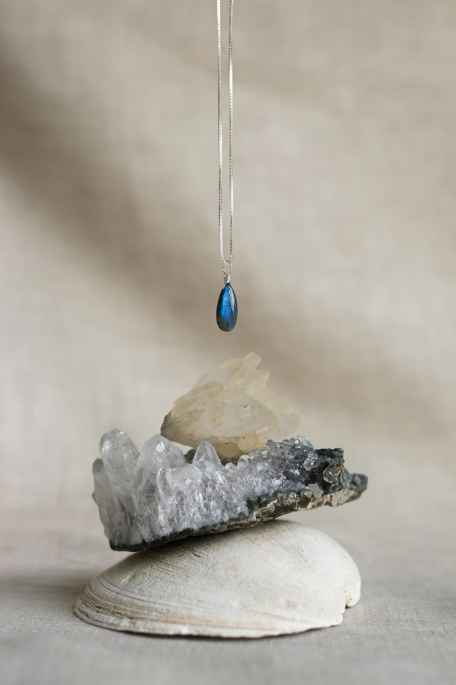 A handcrafted Labradorite teardrop necklace pendant showing a stunning blue and green iridescent flash, hanging above crystals and a shell.