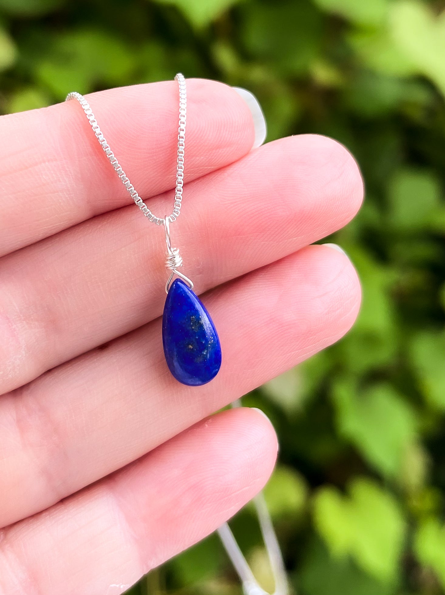 Lapis lazuli teardrop pendant on a silver chain held in a hand with a blurred green background