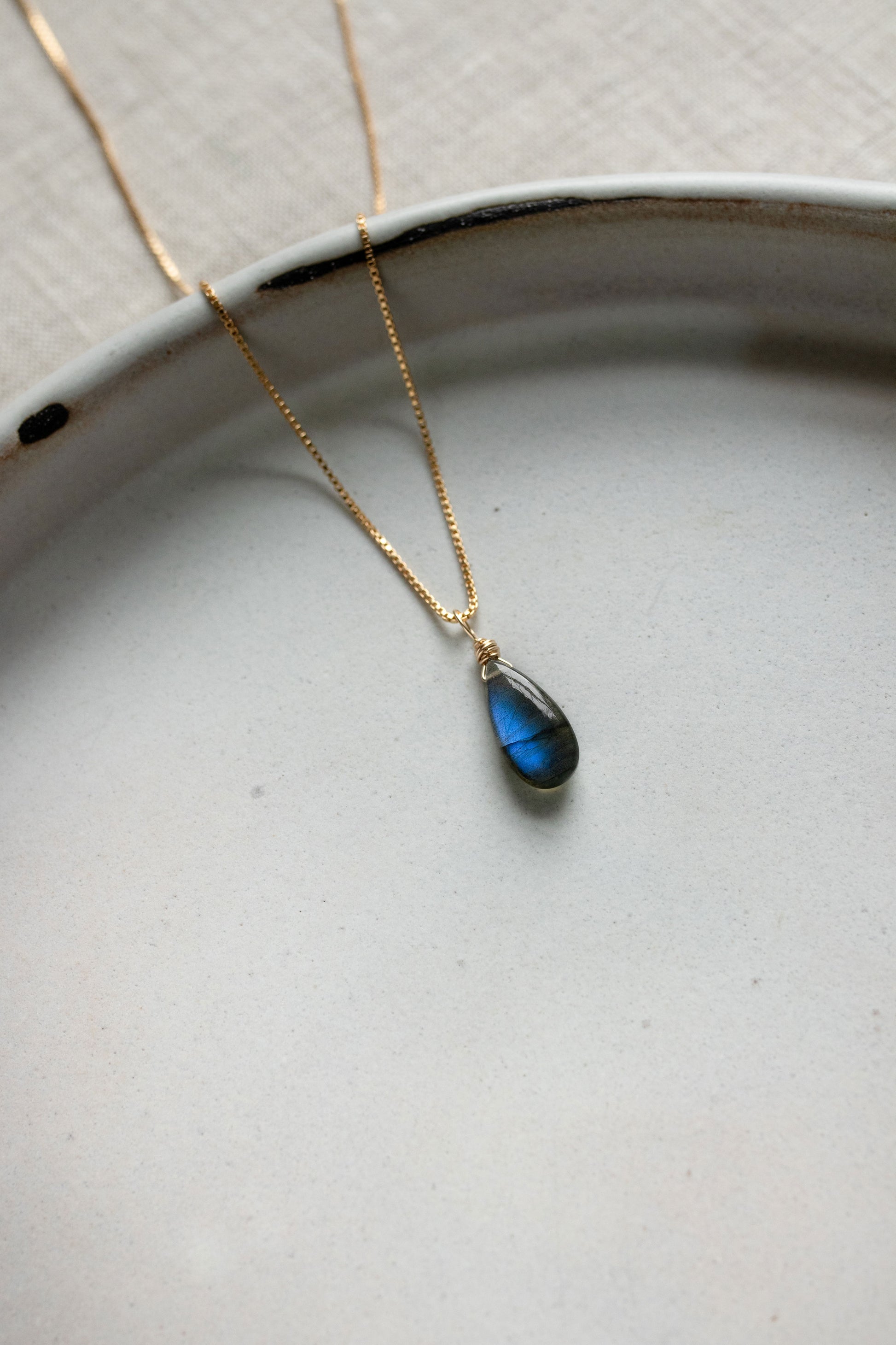A handcrafted Labradorite teardrop necklace pendant showing a stunning blue and green iridescent flash, on a gold chain.