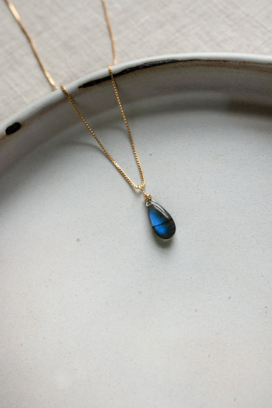 A handcrafted Labradorite teardrop necklace pendant showing a stunning blue and green iridescent flash, on a gold chain.