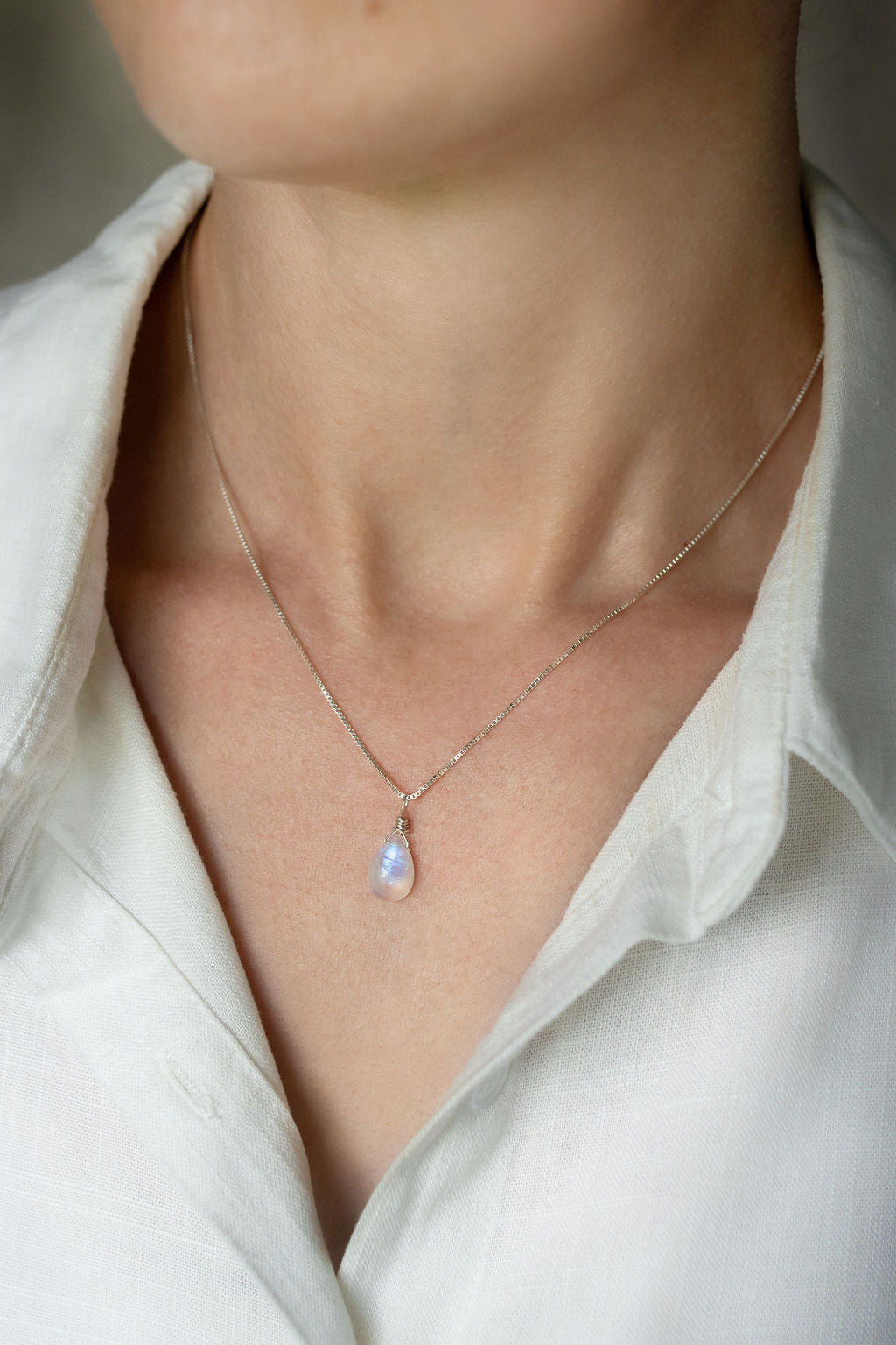 A teardrop-shaped rainbow moonstone necklace with a vibrant iridescent flash, hand wire-wrapped and hanging from a box chain with a spring-ring clasp, on a person wearing a white shirt.