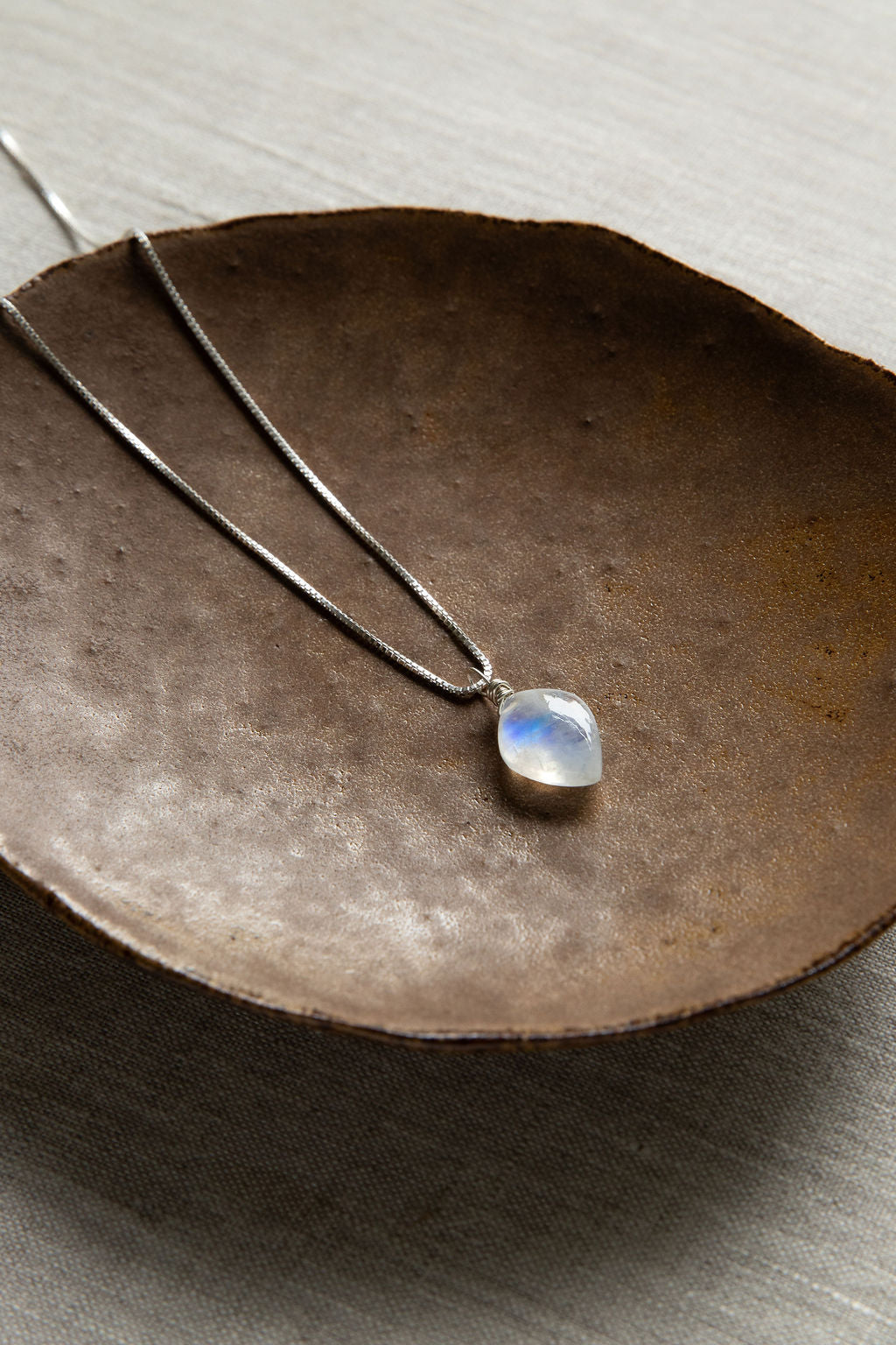 A handcrafted leaf-shaped Rainbow Moonstone necklace with a wire-wrapped crystal pendant on a .925 sterling silver box chain, set on a brown bowl.