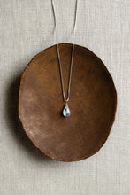 Load image into Gallery viewer, A teardrop-shaped rainbow moonstone necklace with a vibrant iridescent flash, hand wire-wrapped and hanging from a box chain with a spring-ring clasp, set on a brown plate.
