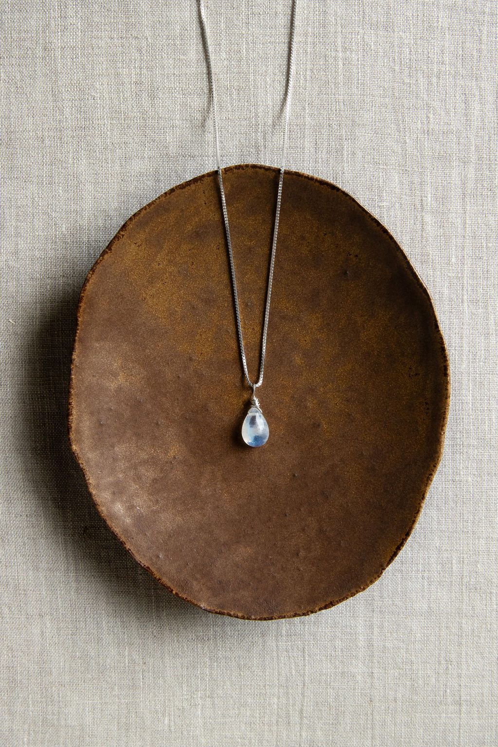 A teardrop-shaped rainbow moonstone necklace with a vibrant iridescent flash, hand wire-wrapped and hanging from a box chain with a spring-ring clasp, set on a brown plate.