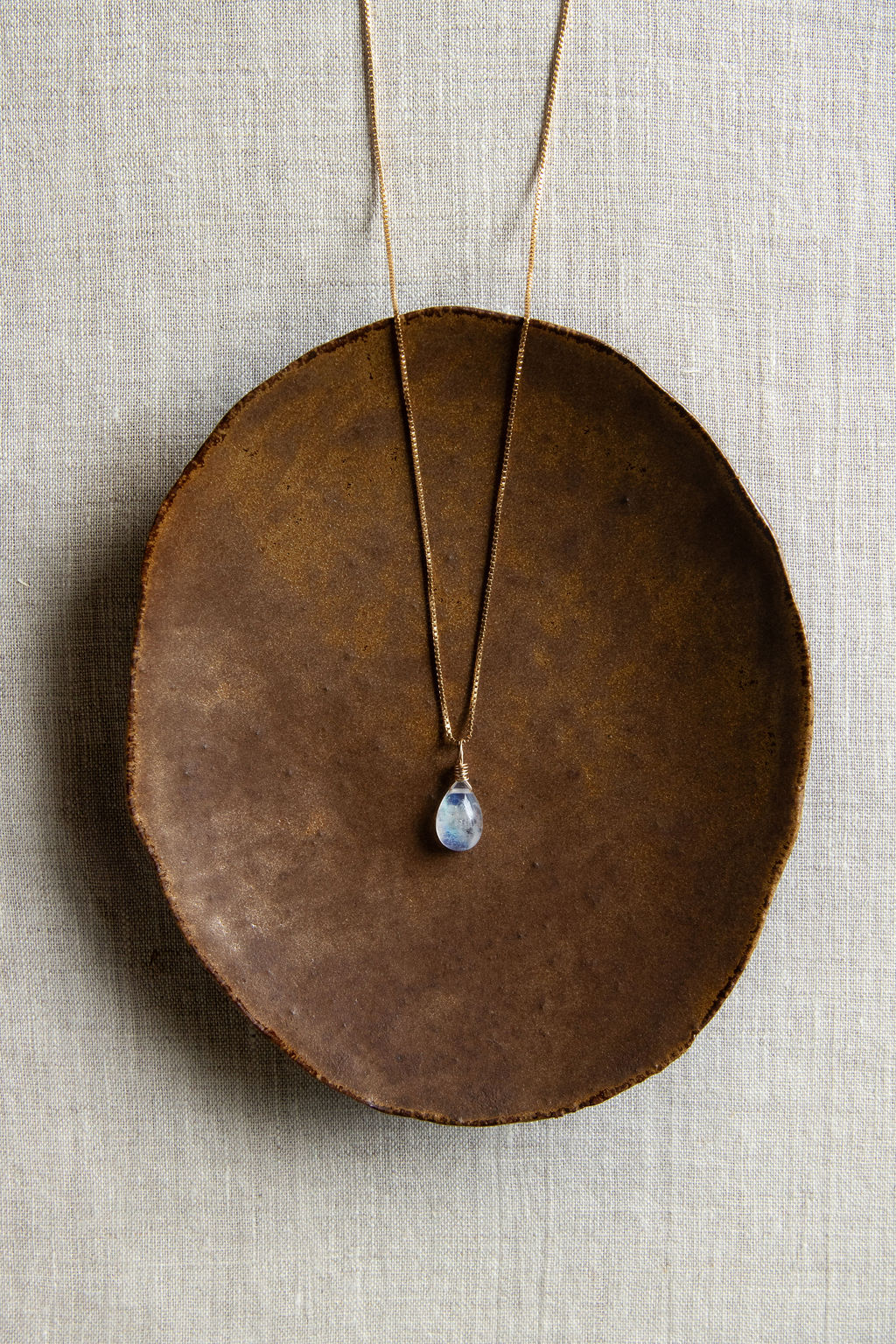 A teardrop-shaped rainbow moonstone necklace with a vibrant iridescent flash, hand wire-wrapped and hanging from a box chain with a spring-ring clasp, set on a brown plate.